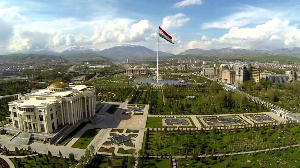 Un paisaje urbano con un edificio destacado de cúpula dorada, un asta con bandera y un parque bien cuidado con jardines geométricos. 1