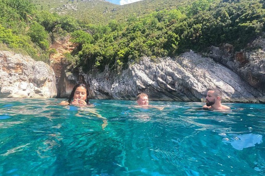 Three people swimming in a clear blue pool with rocky cliffs and greenery in the background. 2