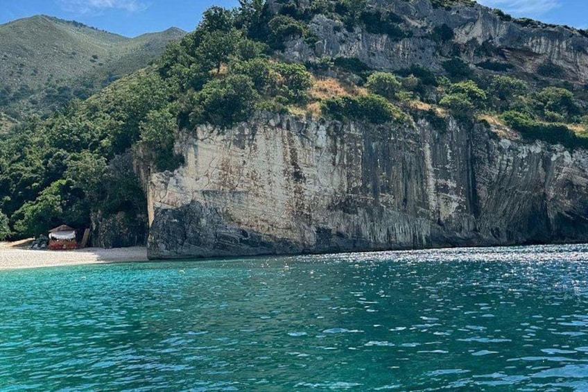 A coastal landscape with a beach, clear blue water, and a rocky cliff with vegetation. 3