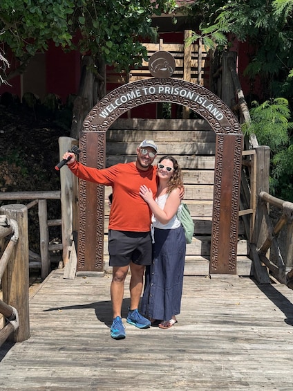A wooden archway with 'Welcome to Prison Island' text, a man in an orange shirt and shorts, and a woman in a white top and blue skirt standing on a wooden deck. 2