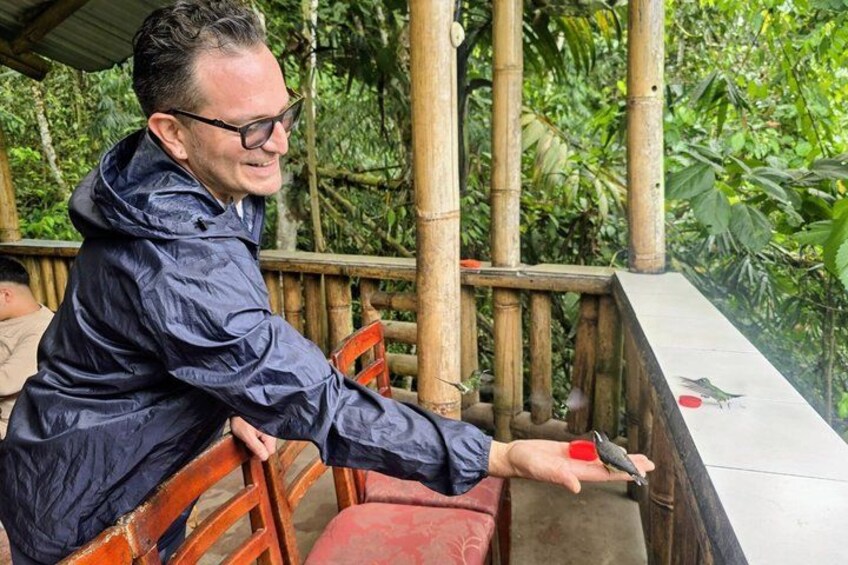 A person in a blue jacket holding a red cup, seated at a wooden table with a bamboo railing in a lush, green environment. 3