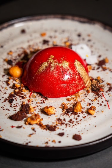 A dessert with a red dome on a plate with scattered crumbs and gold flakes. 5