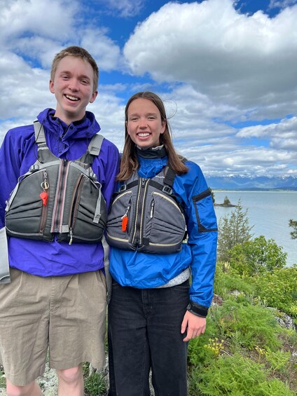 Two people with backpacks standing on a rocky shore with a lake and mountains in the background. 3
