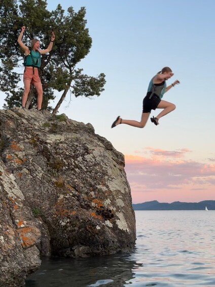 Two people jumping off a rocky cliff into water, with a sailboat in the distance and a sunset sky. 5