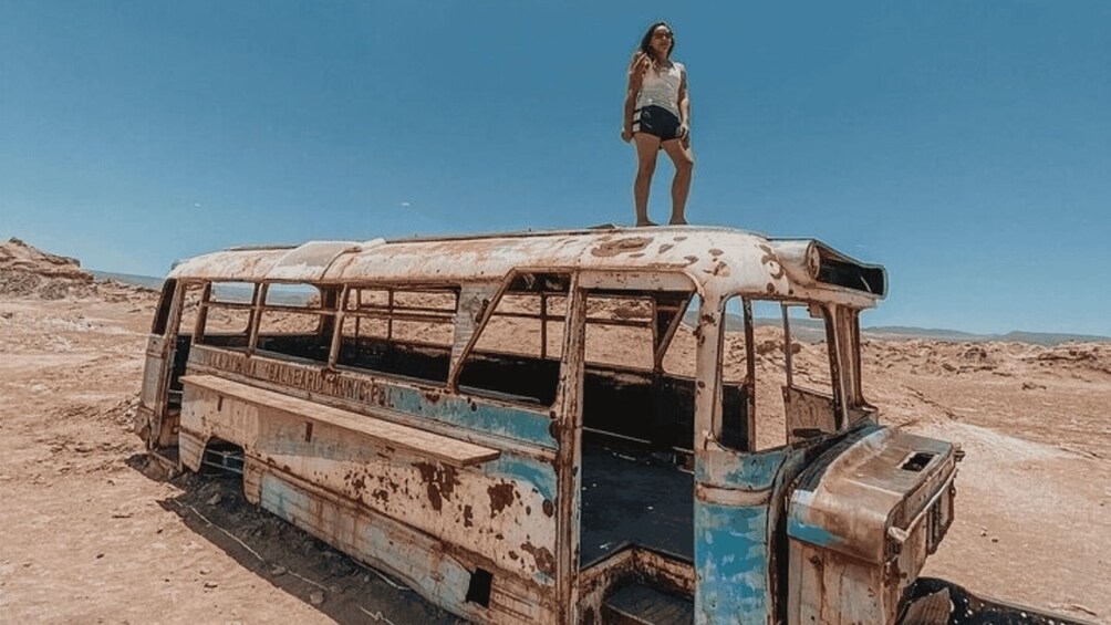 A woman standing on an abandoned, rusted bus in a desert landscape. 3