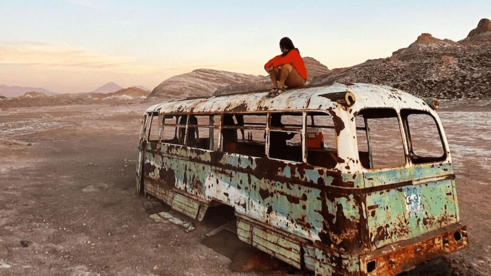 A person sitting on a rusted, abandoned bus in a barren landscape with mountains in the background. 4
