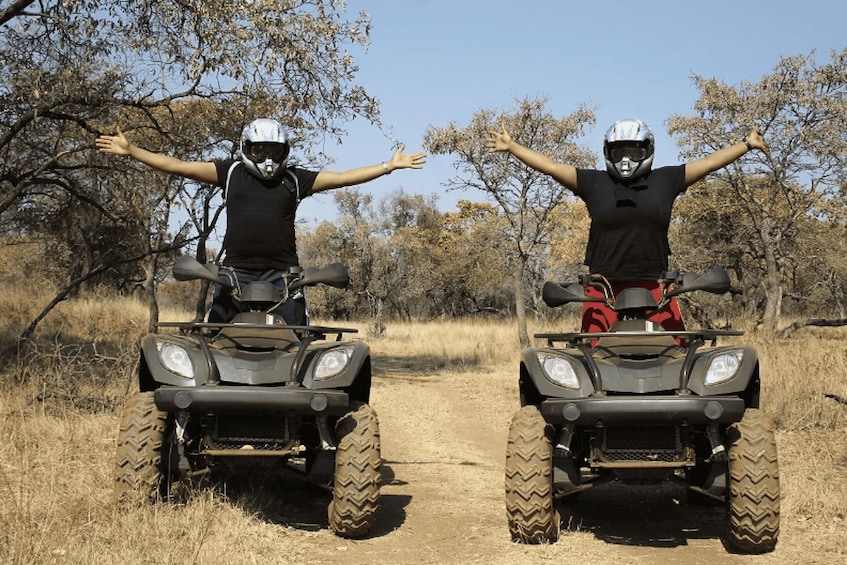 Two people on ATVs with their arms outstretched on a dirt path surrounded by dry grass and trees. 3