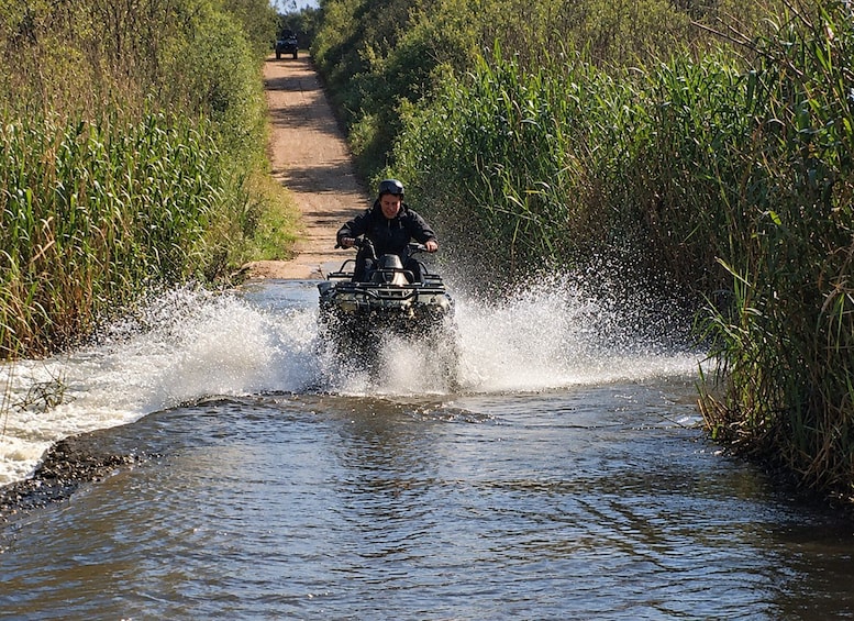 A person riding an ATV through a narrow water channel surrounded by tall vegetation. 4