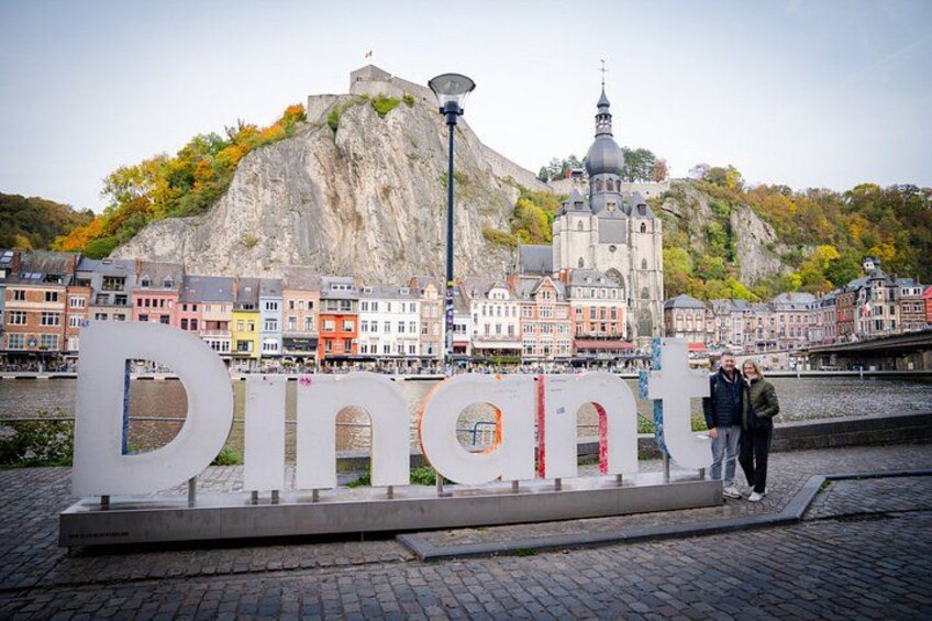 A sign with the word 'Dinant' in large letters, a river, colorful buildings, and a hill with a church. 1