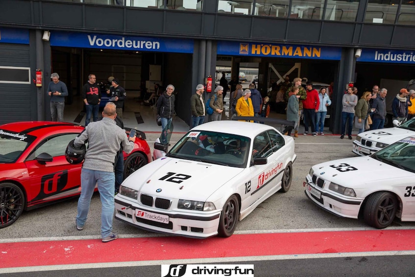 Un grupo de personas frente a un edificio con letreros 'Voordueren' y 'HORMANN', con tres coches BMW blancos estacionados cerca. 2