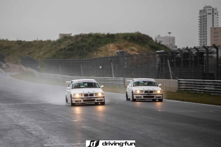 Dos coches de carreras blancos en una pista de carreras mojada, con una valla y edificios al fondo. 5