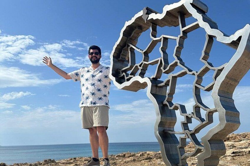 A person standing next to a large metal sculpture on a beach with the ocean in the background. 1