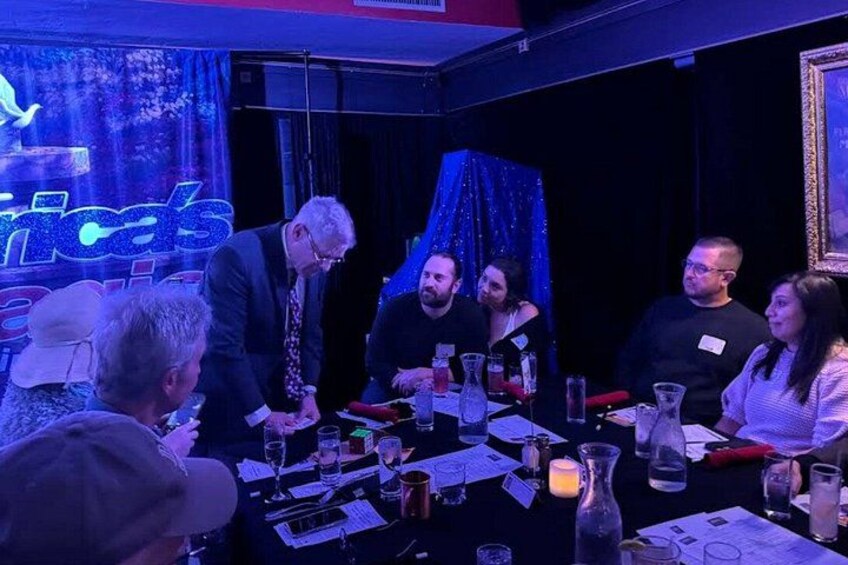 A group of people seated at a table with a blue curtain backdrop, engaged in a discussion or meeting. 3