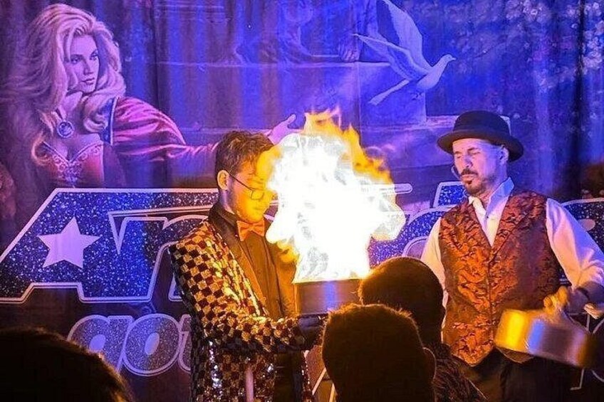 A stage with a performer in a checkered outfit and another in a vest, both with a fire effect in the background. 1