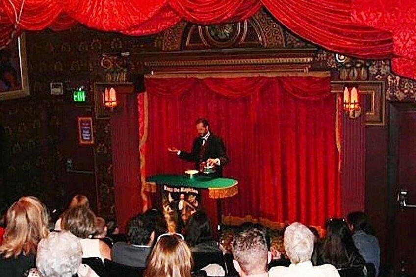 A person speaking at a podium in a theater with red curtains and ornate decorations. 2