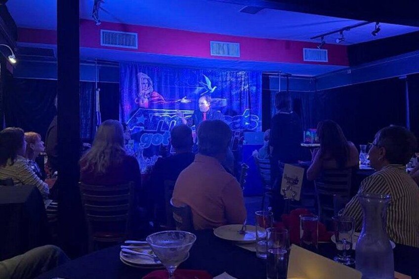A stage with a performer, an audience seated at tables, and a neon sign. 4