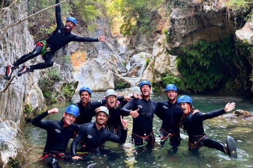 A group of people in wetsuits and helmets near a rocky waterfall. 1