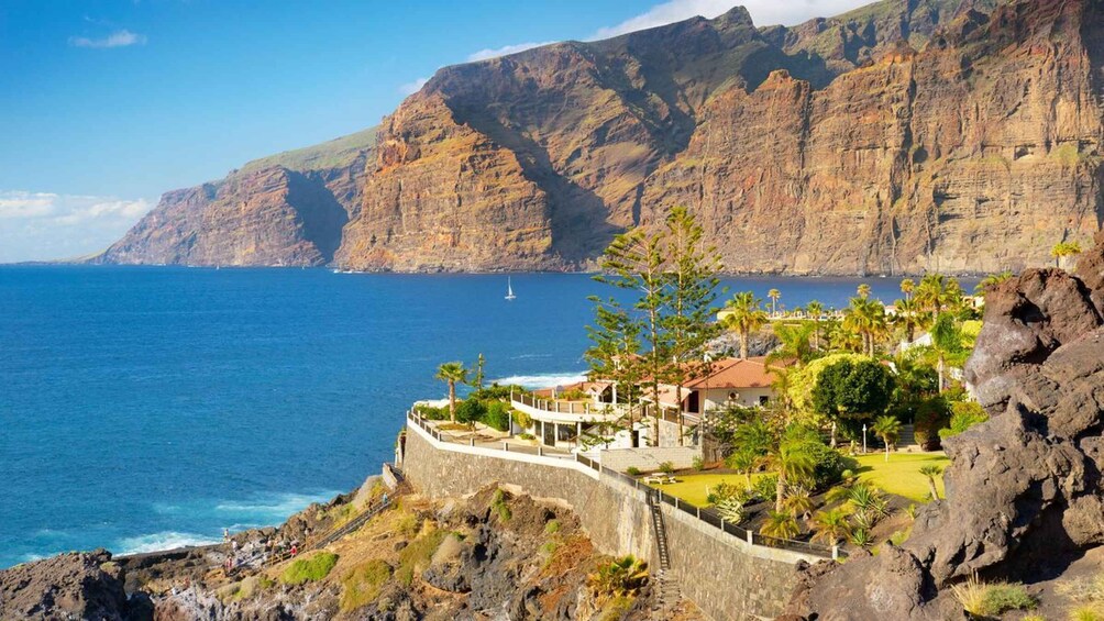 A coastal landscape with a rocky cliff, a clear blue sea, and a small settlement with buildings and vegetation. 1