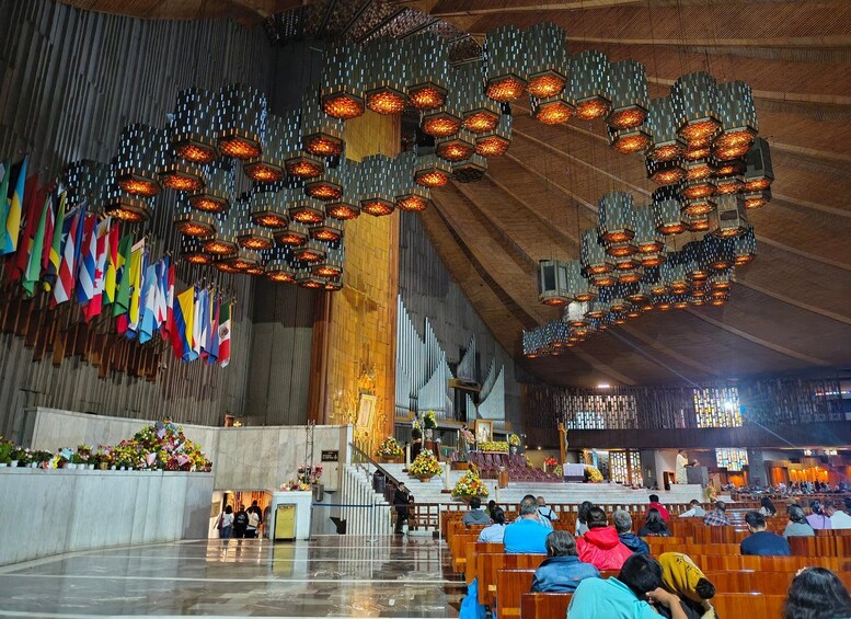 A church interior with wooden pews, a large altar, and a ceiling with hanging light fixtures. Flags are displayed on the walls. 3