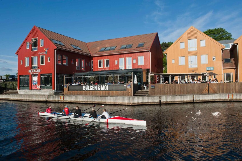 A red building with a sign that reads "BOLGEN & MOI" and a wooden building with a sign that reads "DNB". There is a rowboat with people rowing on the water in front of the buildings. 5