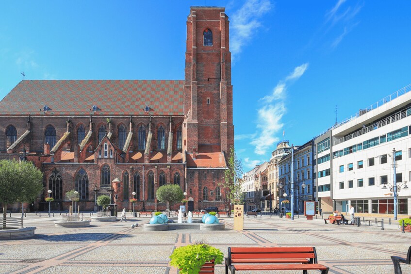 A historic church with a tall tower, a plaza with benches, and a clear blue sky. 2