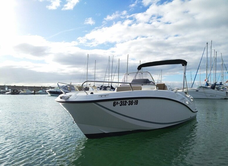 A white motorboat with a registration number, docked at a marina with other boats and masts in the background. 1