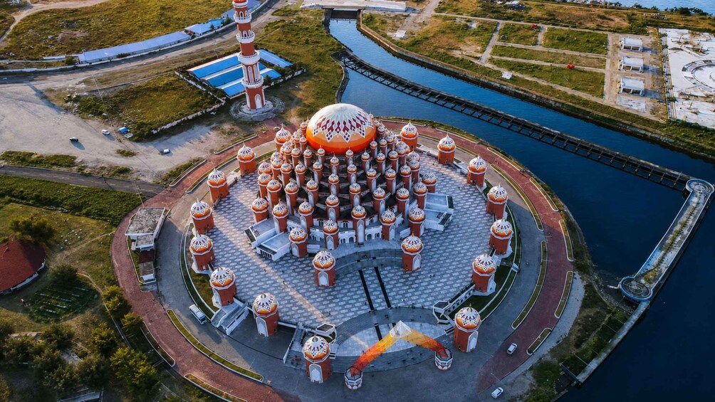 An aerial view of a circular architectural structure with a central dome and multiple smaller domes, surrounded by a paved area and a body of water. 2