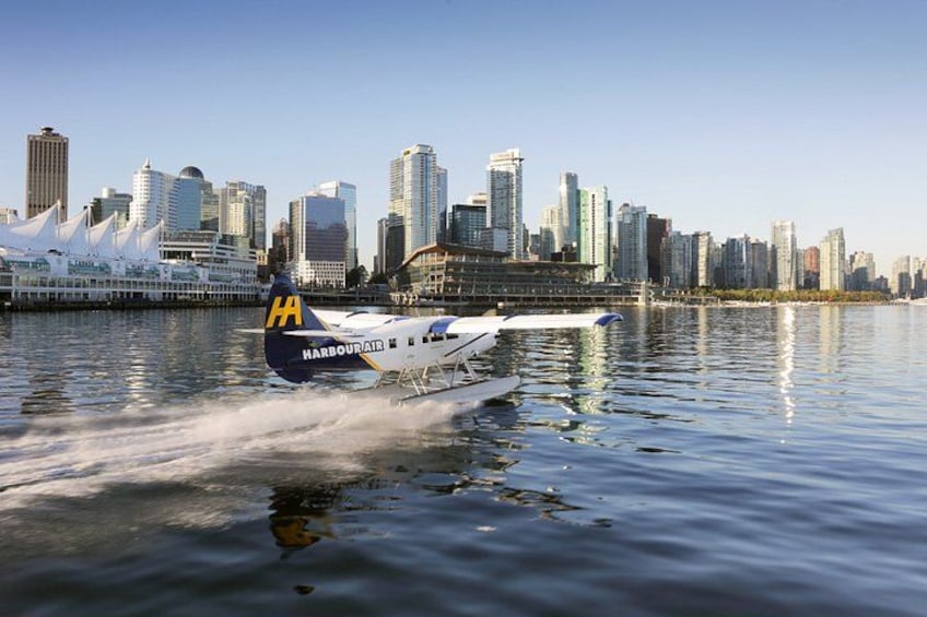 Un hidroavión de HarbourAir con una decoración blanca y azul vuela a baja altura sobre una masa de agua, con un horizonte urbano de fondo. 3