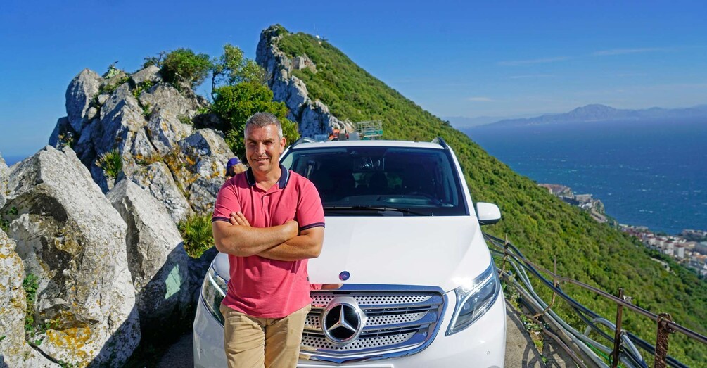 A man standing next to a white Mercedes-Benz car on a mountain road with rocky terrain and a view of the sea in the background. 1