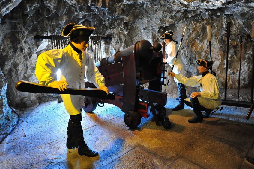 A historical reenactment scene with actors in period costumes, a cannon, and a cart in a rocky cave setting. 2