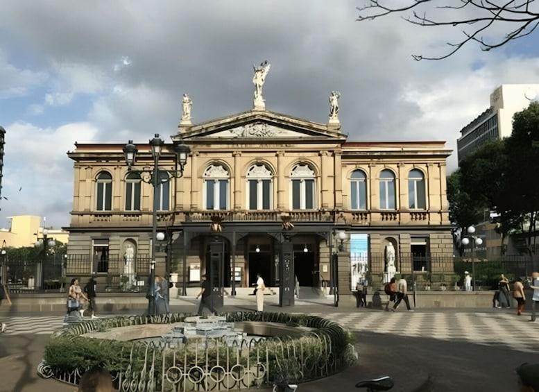A historic building with statues on the roof, a central fountain, and people walking around. 2