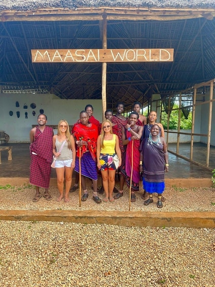 A group of people posing in front of a structure labeled "MAASAI WORLD" with a thatched roof and wooden beams. 1