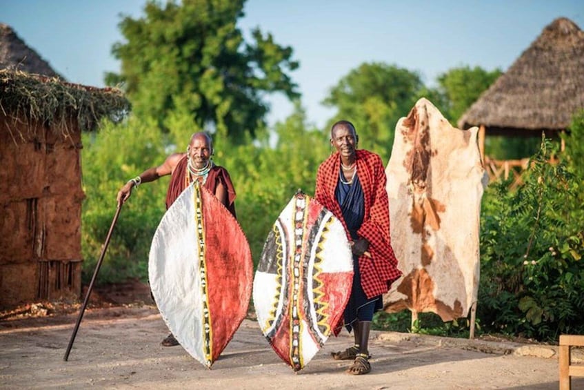 Two people carrying large shields, walking past traditional huts in a rural setting. 4