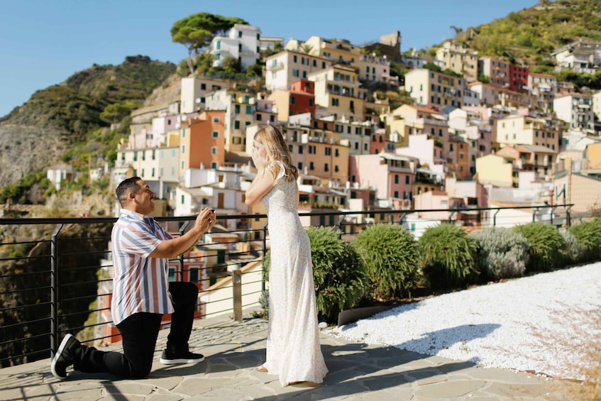 Un homme à genoux, demandant une femme en mariage alors qu’elle porte une robe blanche, sur un chemin de pierre avec des bâtiments colorés en arrière-plan. 5