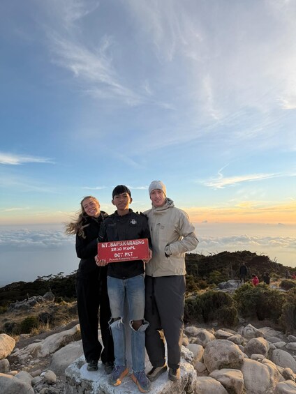 Three people standing on a rocky outcrop with a sign in front of them. 2