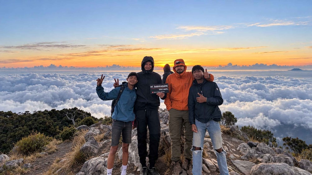 Four people standing on a rocky mountain peak with a view of clouds below and a sunset in the background. 1