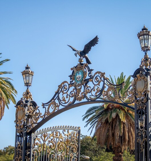 An ornate wrought iron gate with a bird sculpture, two street lamps, and palm trees in the background. 4