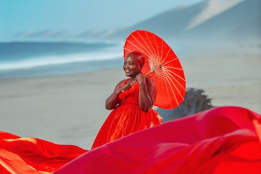 Eine Person in einem roten Kleid hält einen roten Regenschirm am Strand, im Hintergrund Dünen. 5