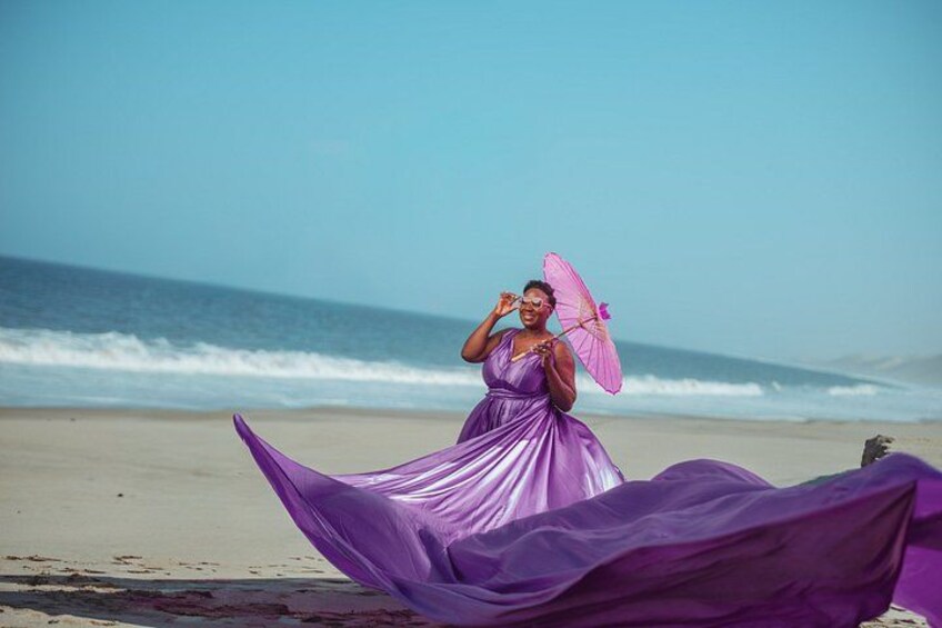 Eine Person in einem wallenden violetten Kleid steht an einem Strand, im Hintergrund sind Wellen zu sehen. 3