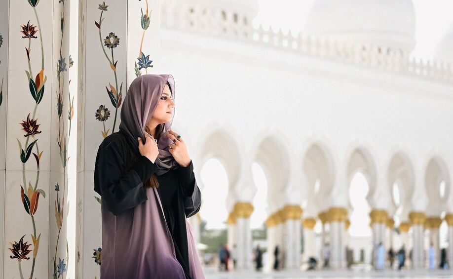 A person in a purple dress and black jacket standing in a white building with arches. 3