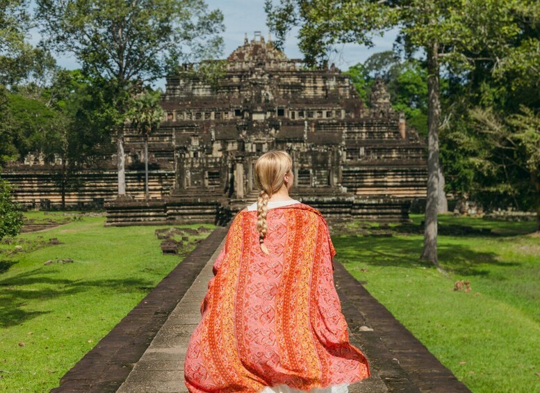 A person in a red patterned garment walking on a stone pathway towards a large, ancient temple surrounded by trees. 1