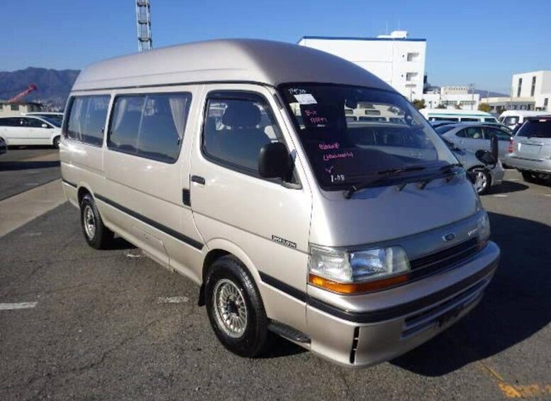 A silver van parked in a lot with other vehicles, buildings, and mountains in the background. 3