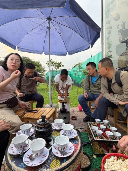 A group of people gathered around a table with cups and a kettle, under a blue umbrella. 2