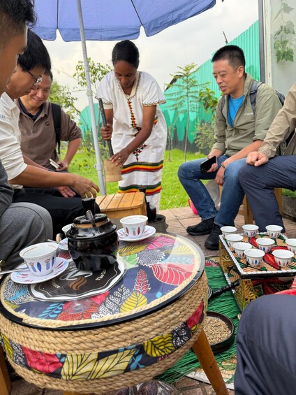 A group of people sitting outdoors under a blue umbrella, with a table displaying cups and a coffee pot. 5