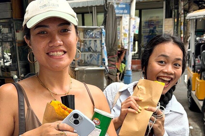Two people holding food items and a drink, standing in a street with shops and a yellow vehicle in the background. 3
