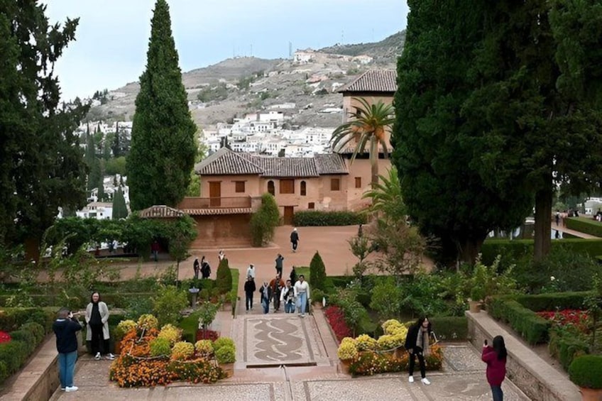 A garden with people, a building with a tiled roof, and a hill with houses in the background. 4