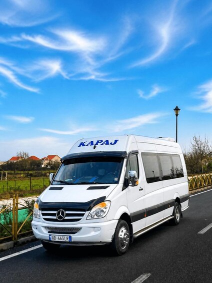 A white van with 'KAPAJ' on the front, parked on a road with a clear blue sky and a lamppost in the background. 3