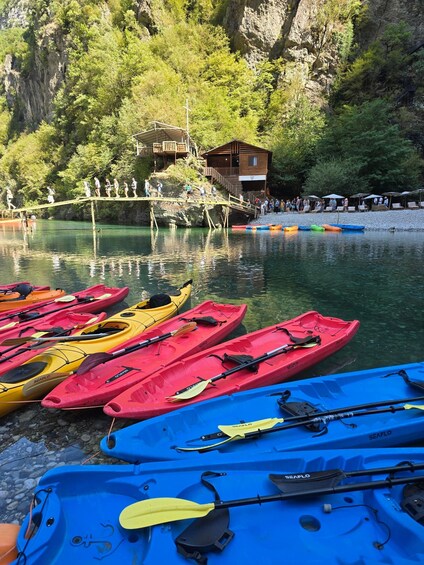 A calm lake with kayaks lined up on the shore, a wooden cabin, and a rocky cliff in the background. 2