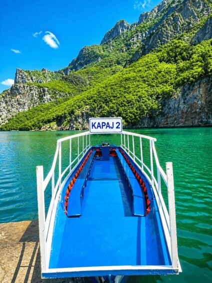 A boat with a blue deck and a sign reading 'KAPAJ 2' on a calm body of water surrounded by green hills. 2