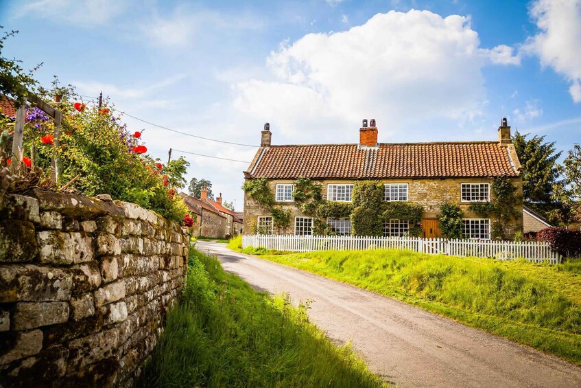A rural road with a stone wall, a white picket fence, and a stone house with a tiled roof. 1